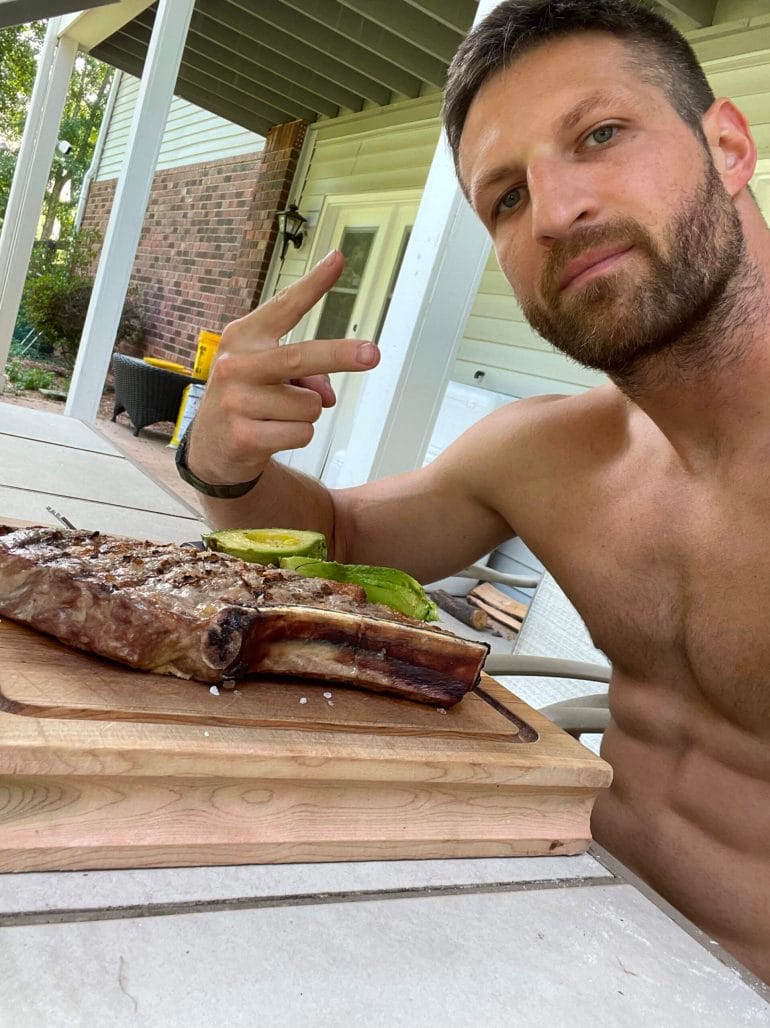 Michael Kummer shirtless and eating a giant piece of meat with an avocado.